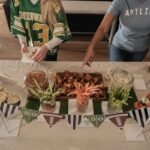 Tailgating party food ideas spread with wings, pretzel jars, popcorn cups and veggie sticks on a football themed table