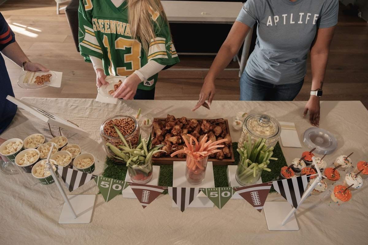 Tailgating party food ideas spread with wings, pretzel jars, popcorn cups and veggie sticks on a football themed table