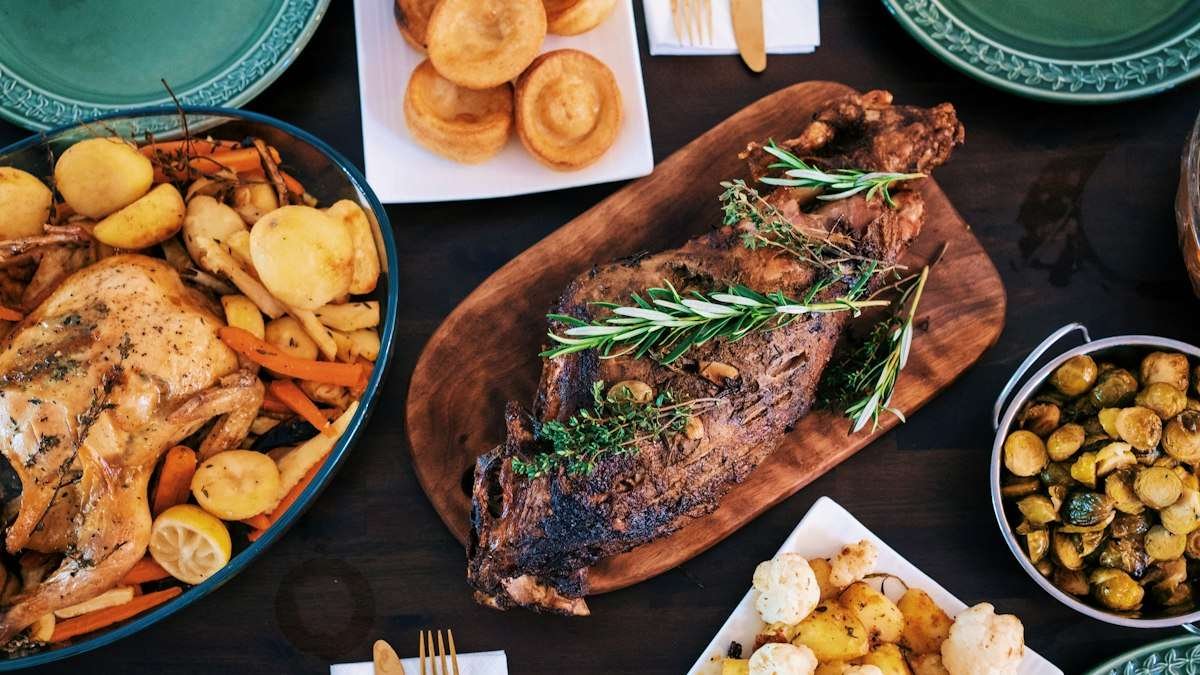 Thanksgiving side dishes spread across a dark table: rolls, roasted vegetables, Brussels sprouts and potatoes around the roast